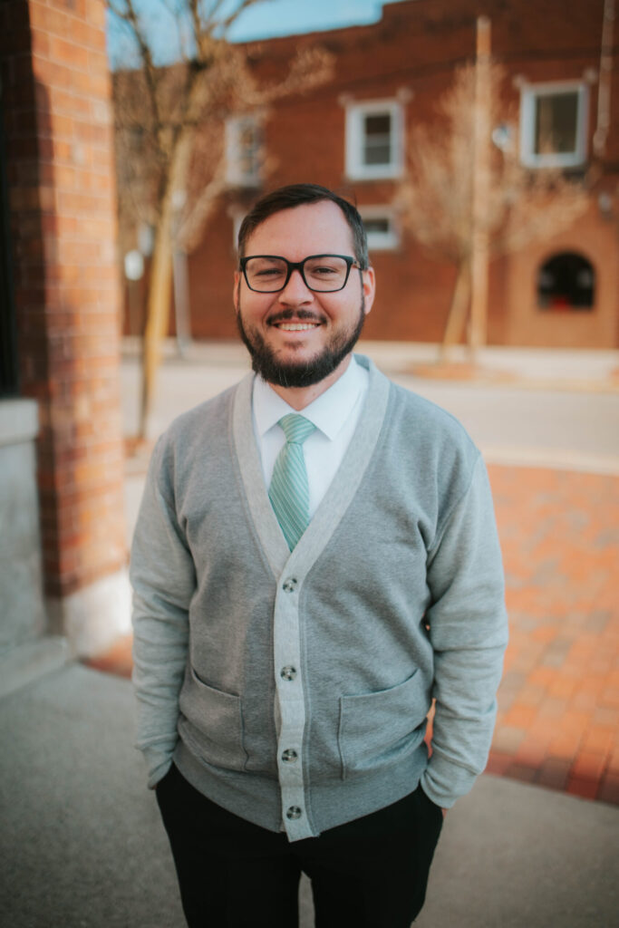 A man with glasses and a beard, wearing a gray cardigan, white shirt, and light green tie, stands smiling outdoors on a brick sidewalk with buildings and bare trees in the background.