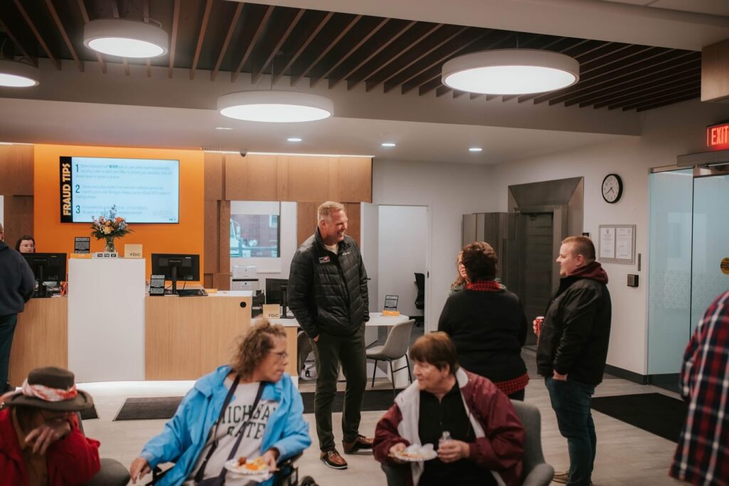 A group of people are socializing in a modern office lobby with wood accents, bright ceiling lights, and a reception desk in the background. Some are seated, while others stand and converse near the center.