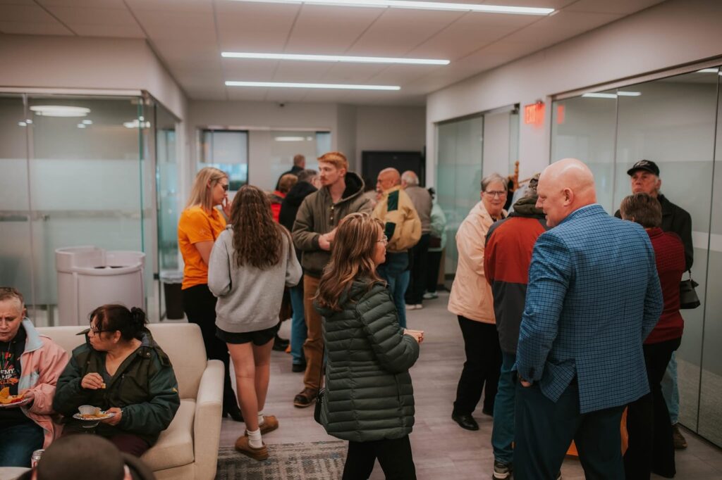 A group of people gather and mingle in a modern office hallway, some standing and chatting, others sitting and eating, creating a lively and social atmosphere.