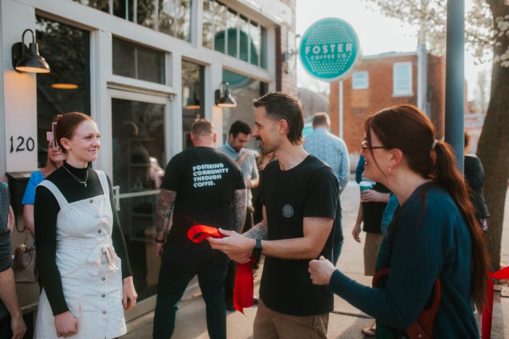 A group of people stand and smile outside Foster Coffee Co. One person holds a red ribbon, and others chat near the entrance, with a green Foster Coffee Co. sign visible above the door.