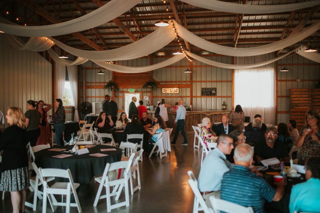 A group of people socialize and dine at round tables in a spacious, rustic barn decorated with white drapes and string lights. Some guests are seated, while others stand and mingle near the back.