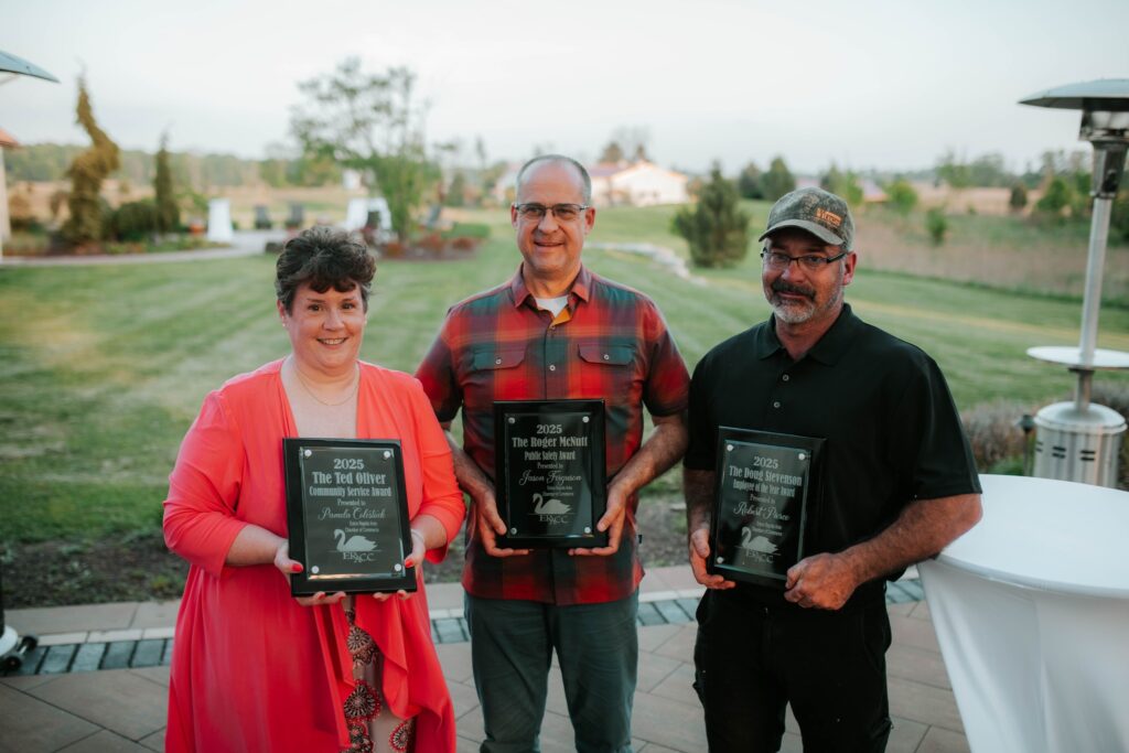 Three people standing outdoors, each holding a black award plaque and smiling at the camera. The background features a grassy lawn, trees, and a walkway on a clear day.