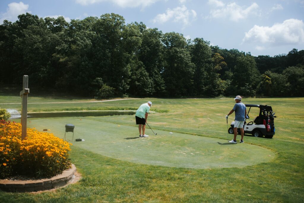 Two people are on a golf course; one is preparing to putt while the other stands nearby with a golf club. A golf cart and flowers are visible, with trees and blue sky in the background.