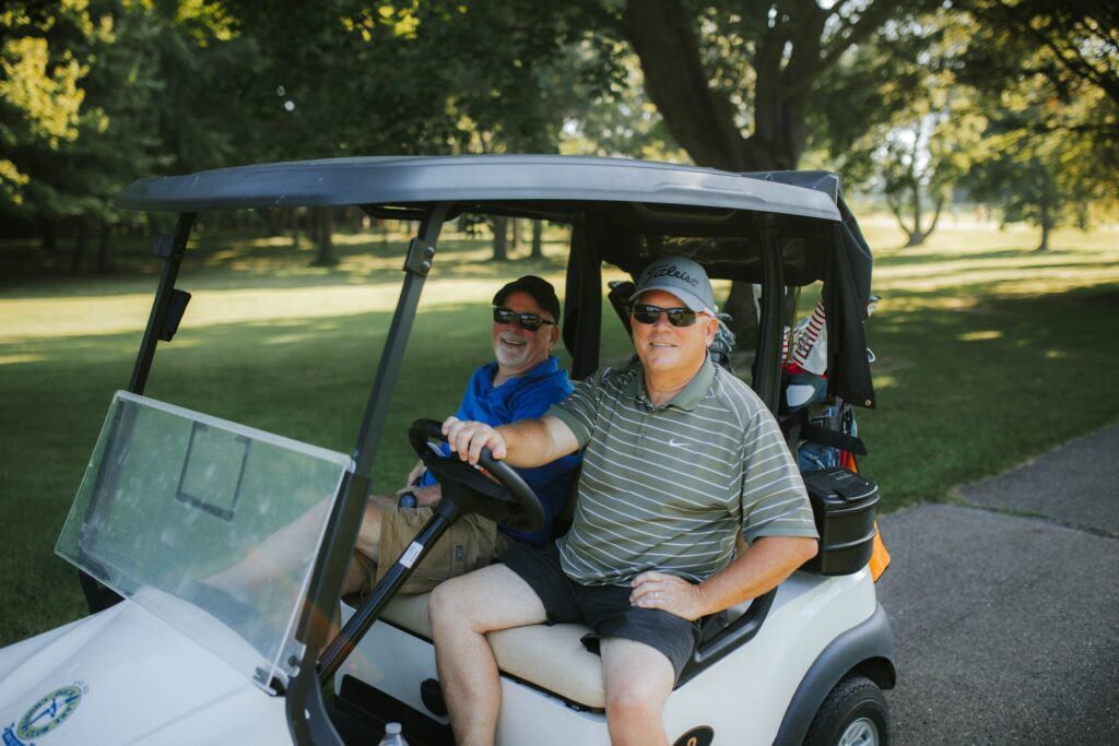 Two men wearing sunglasses and casual clothes sit in a golf cart on a sunny golf course, smiling at the camera with green trees and grass visible in the background.