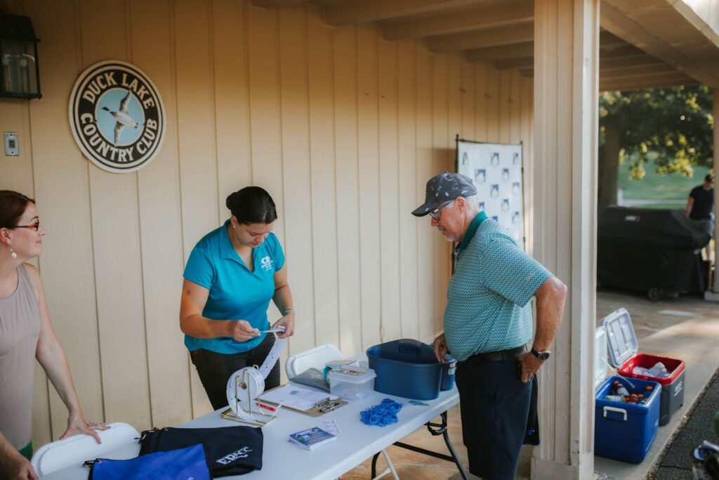 A woman in a blue shirt assists an older man at a check-in table outside Duck Lake Country Club. The table has papers, lanyards, and supplies; another woman stands nearby. A sign and coolers are visible in the background.