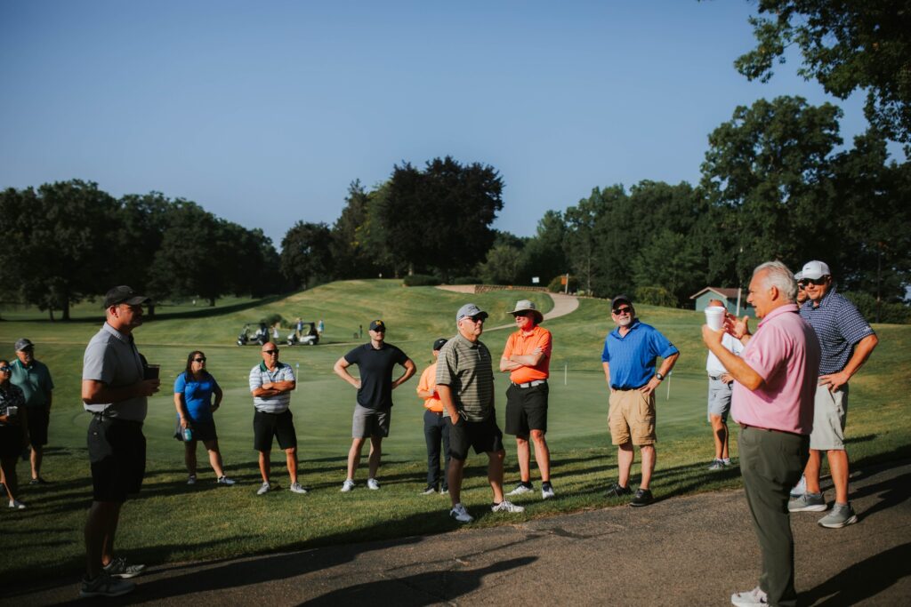 A group of people stands on a golf course, listening to a man in a pink shirt speaking. Golf clubs and carts are in the background, and trees and grass surround the scene on a sunny day.