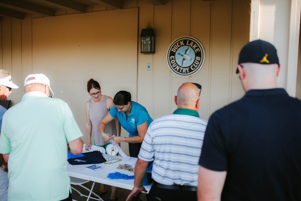 Two women stand behind a table with shirts and papers, assisting four men at an outdoor event near a Duck Lake Country Club sign on a beige wall.