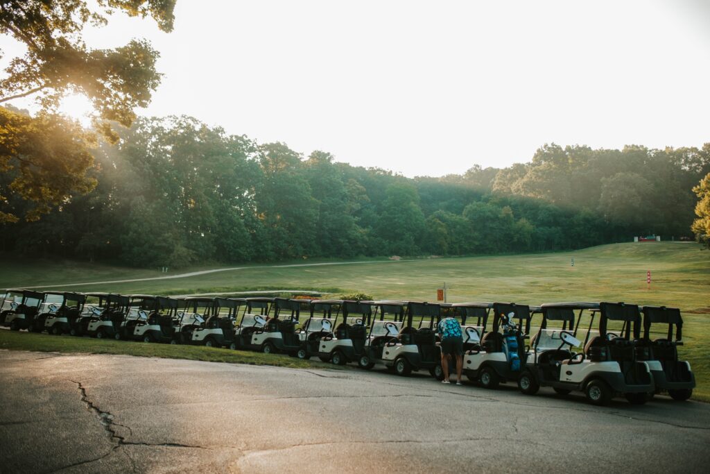 A row of parked golf carts lines the edge of a golf course at sunrise, with two people standing near the carts and trees in the background.