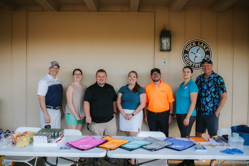 Seven people stand and smile behind a table with colorful towels and snacks displayed. They are under a shelter at Duck Lake Country Club, as shown by the sign on the wall behind them.