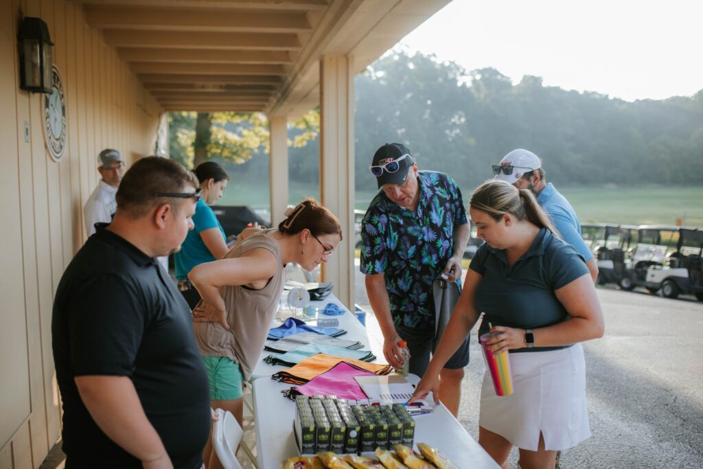 A group of people stand around a table covered with bags, snacks, and papers at an outdoor event near golf carts, with trees and morning sunlight in the background.