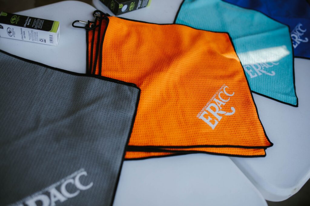 Several microfiber towels in gray, orange, blue, and teal lay on a white surface, each printed with the white text ERICC in bold letters. A small box is partially visible in the top left corner.