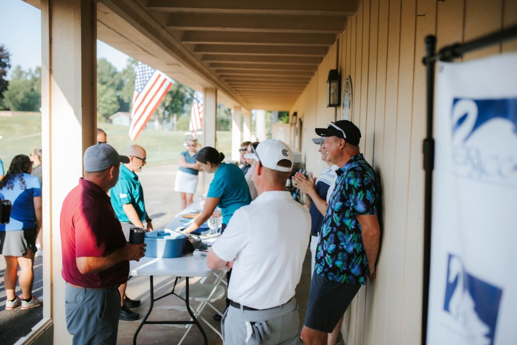 People gather and socialize around a check-in table under a covered walkway at an outdoor event. Some are wearing golf attire, and two American flags are visible in the background.