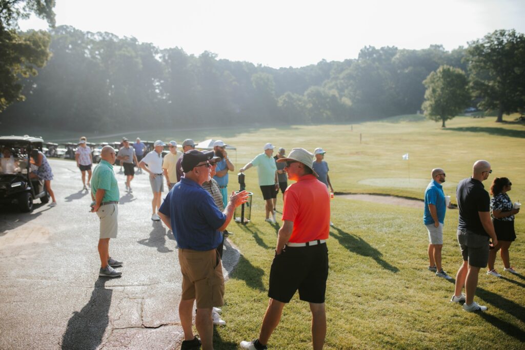 A group of people, mostly men, stand and talk on a sunny golf course near golf carts. Some wear hats and sunglasses, and the area is surrounded by trees and green grass.