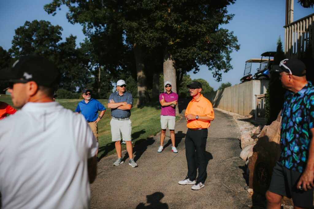 A group of men in casual golf attire stand on a golf course path, listening to a man in an orange shirt speaking. Golf carts and trees are visible in the background on a sunny day.