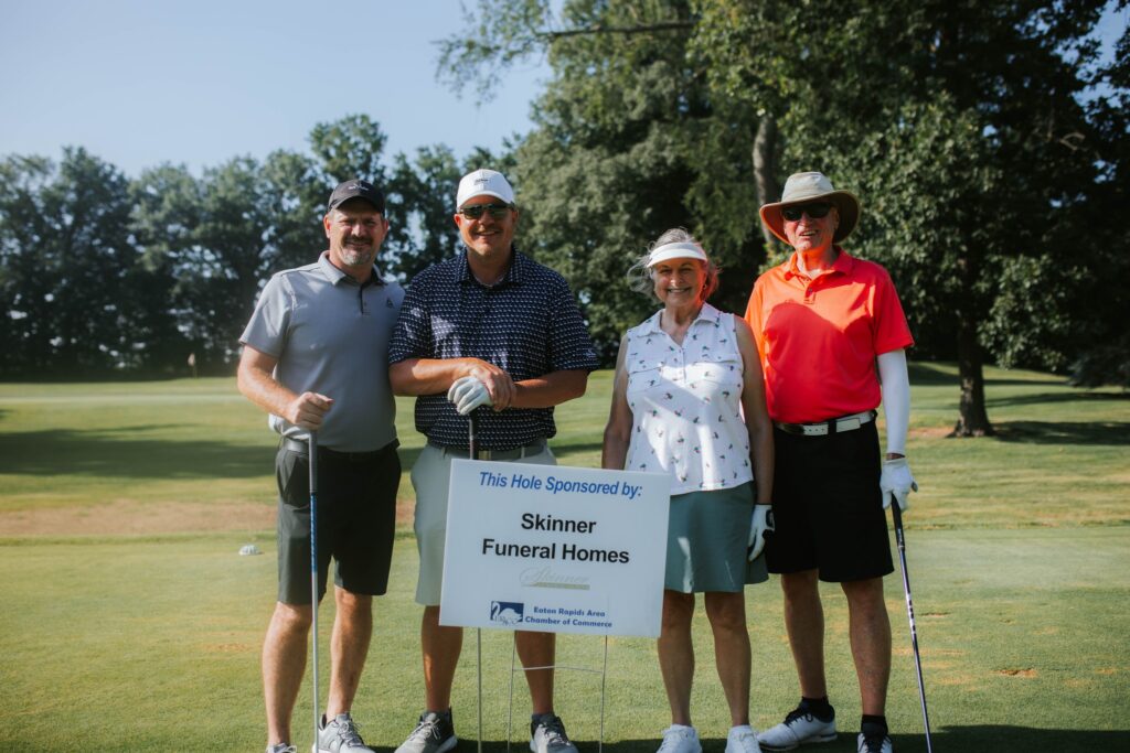 Four people stand on a sunny golf course next to a sign that reads, This Hole Sponsored by: Skinner Funeral Homes. They are smiling and dressed in golf attire, holding clubs, with trees and green grass in the background.