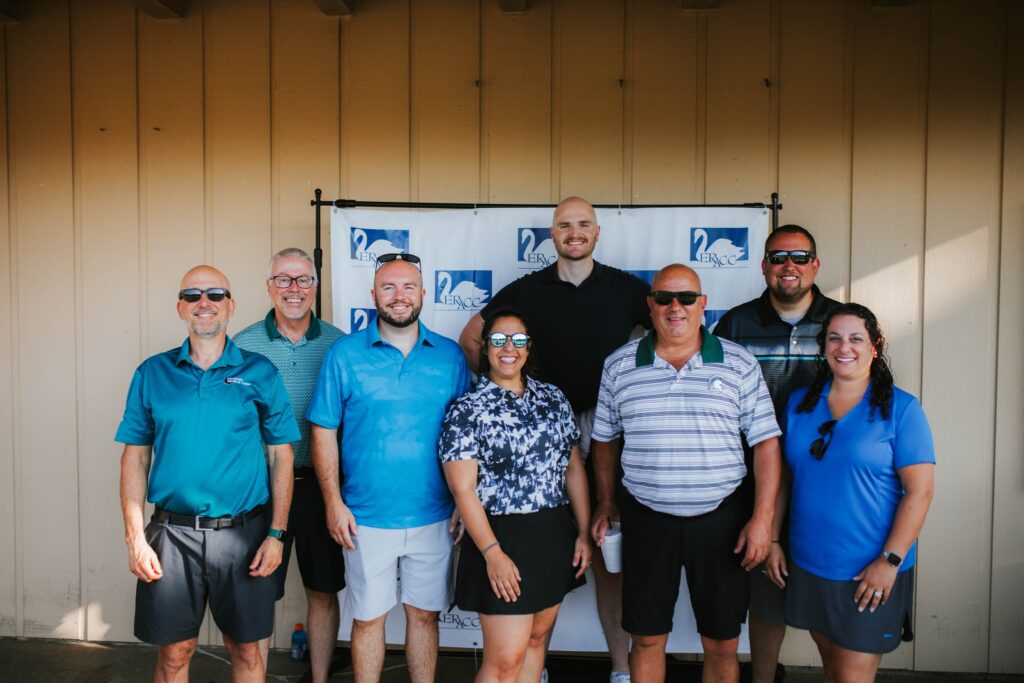 Eight people, five men and three women, stand smiling in front of a beige wall and a banner with blue logos. They are dressed in casual golf attire and sunglasses, posing for a group photo outdoors.