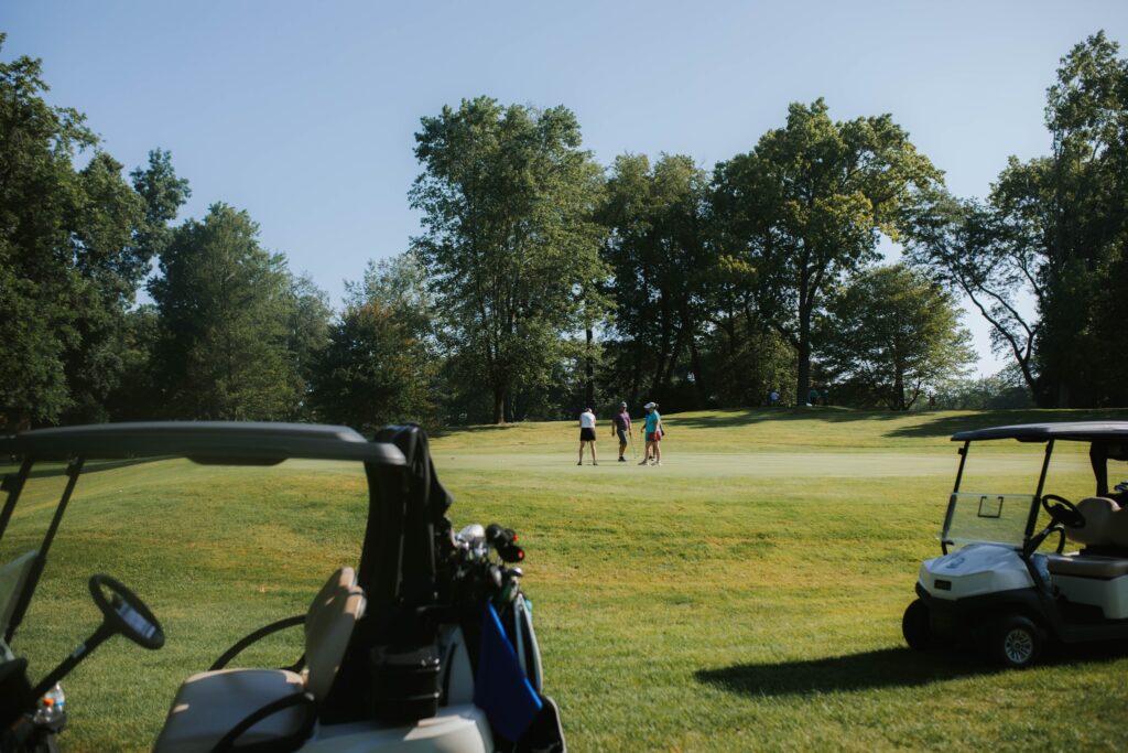 Two people stand on a grassy golf course surrounded by trees, with golf carts parked in the foreground under a clear blue sky.