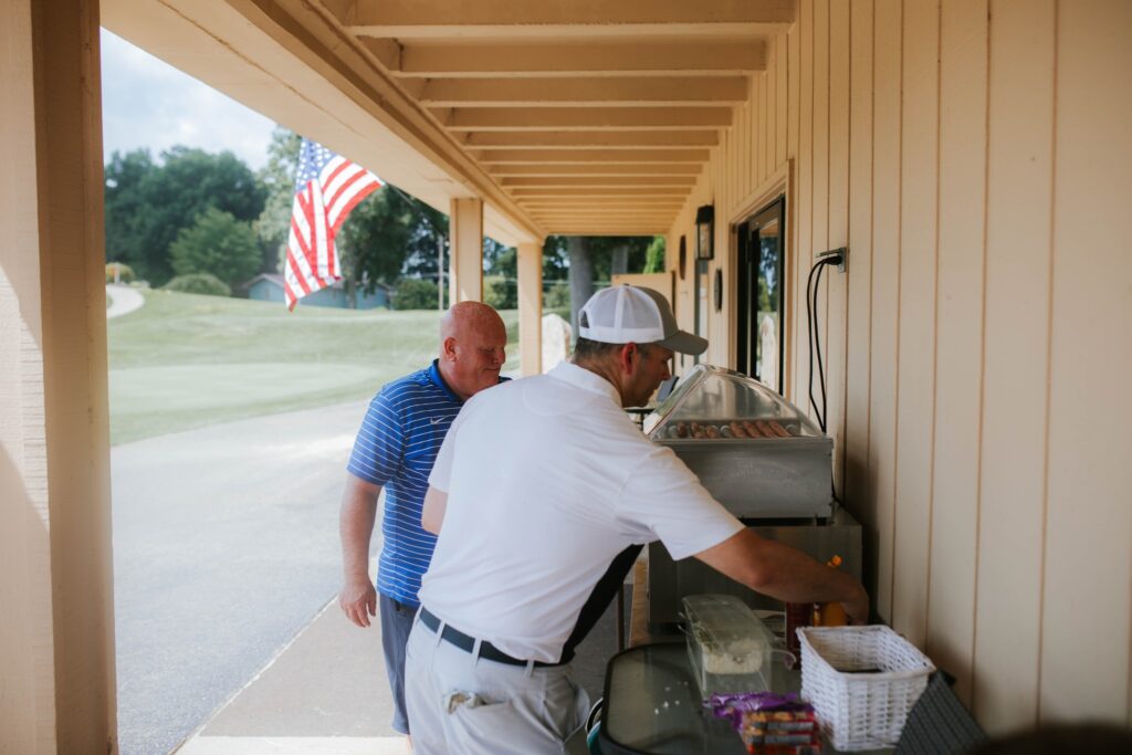 Two men stand at an outdoor snack stand beside a beige building. One man wears a white golf outfit and cap, reaching for snacks, while the other in a blue polo shirt observes. An American flag hangs nearby.