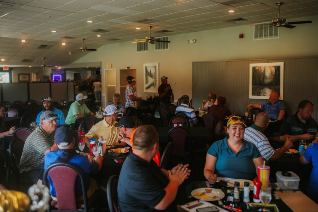 A group of people, mostly men, sit at tables in a large, well-lit dining room, eating, talking, and socializing. The atmosphere is casual and lively, with paintings on the walls and ceiling fans above.