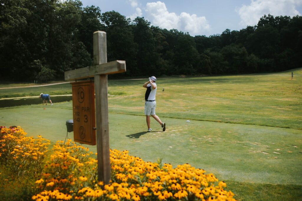 A golfer swings on a bright green golf course near a patch of yellow flowers and a wooden sign reading “10 Par 3.” Dense trees and blue sky with clouds are visible in the background.