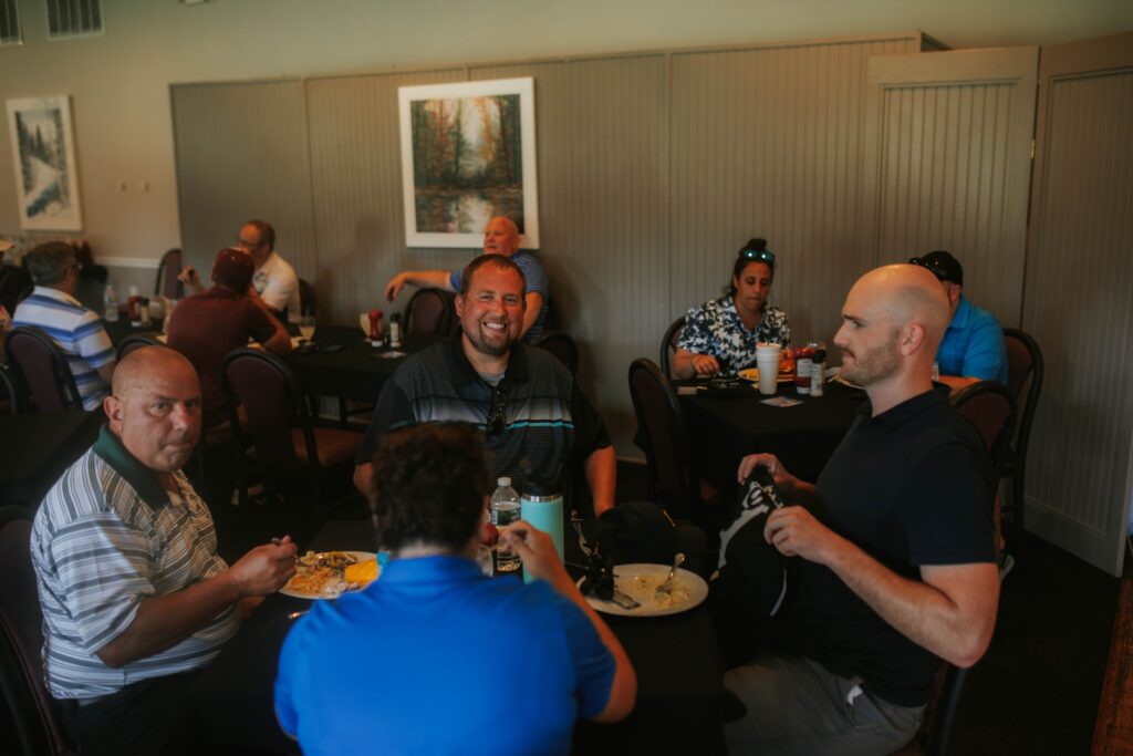 A group of people sit around tables in a restaurant, eating and talking. One person at the center is smiling at the camera. The room has neutral walls and framed art. Plates of food and drinks are on the tables.