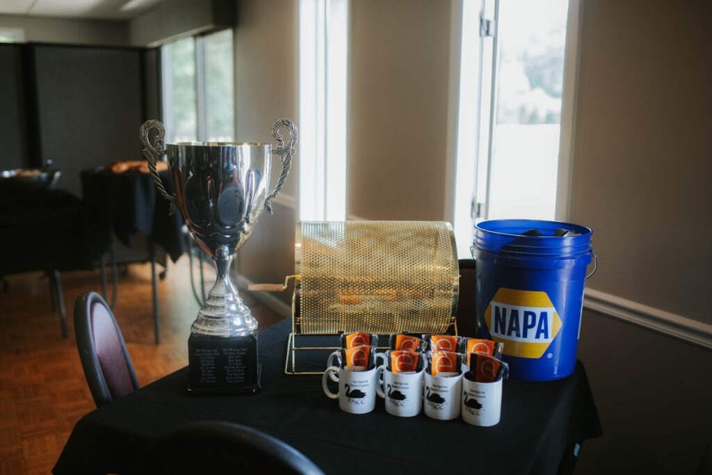A large trophy, a gold raffle drum, a blue NAPA bucket, and coffee mugs with orange cards are displayed on a black tablecloth in a sunlit room with chairs and windows in the background.