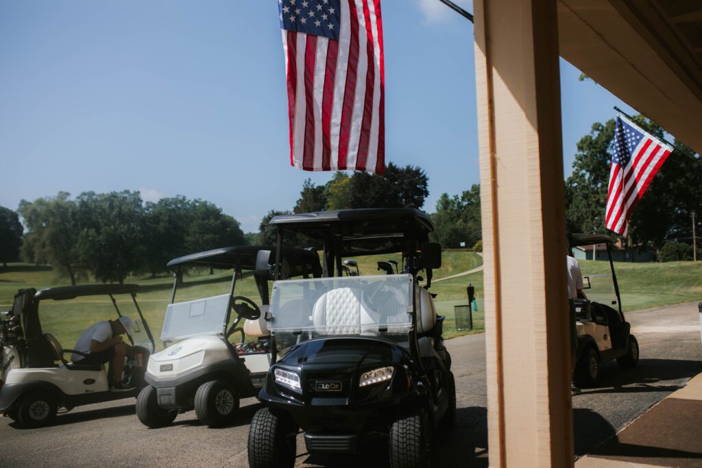 Three golf carts parked outside on a sunny day with large American flags hanging above them. Green grass and trees are visible in the background, creating a scenic golf course setting.