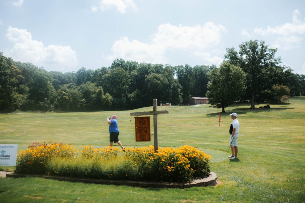 Two people are playing golf on a grassy course surrounded by trees. One person is swinging a club near a wooden golf hole sign, while the other watches nearby. Yellow flowers border the tee area.