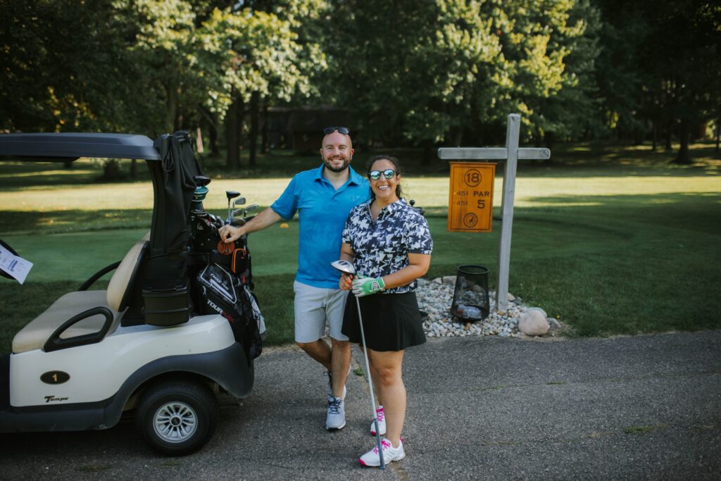 A man and a woman stand smiling next to a golf cart on a golf course near a sign for hole 18, par 5. Both wear golf attire and hold clubs, with trees and grass visible in the background.