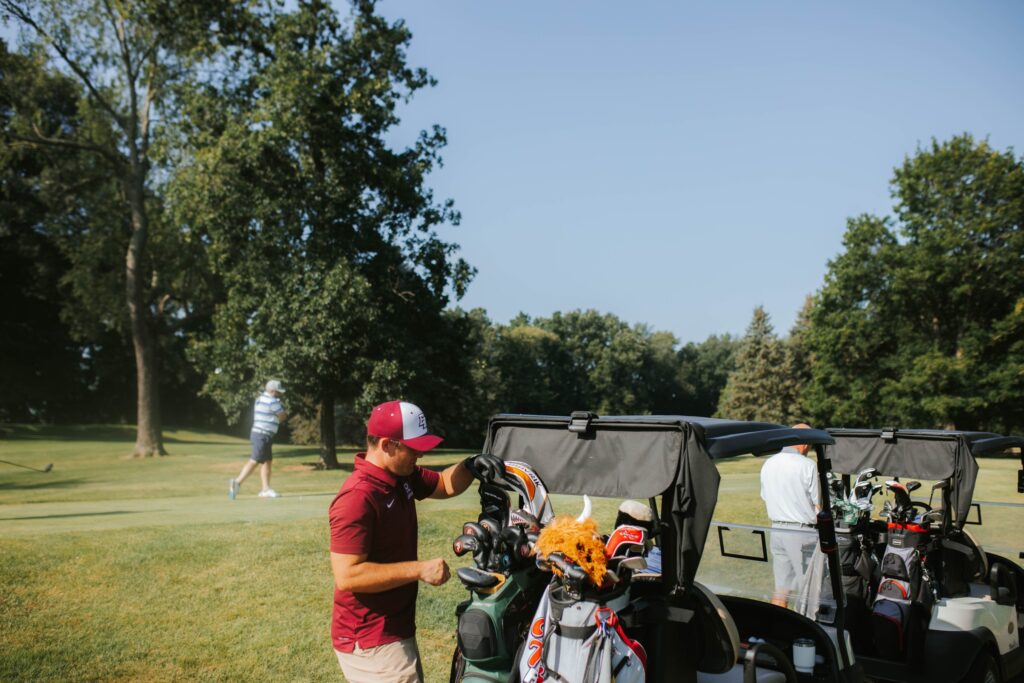 A man in a red shirt and cap stands by a golf cart organizing golf clubs, while two people walk on the green golf course in the background surrounded by trees under a clear sky.