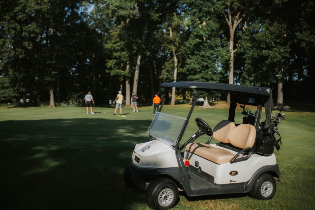 A white golf cart parked on grass in the foreground, with four people standing on a green in the background surrounded by tall trees on a sunny day.