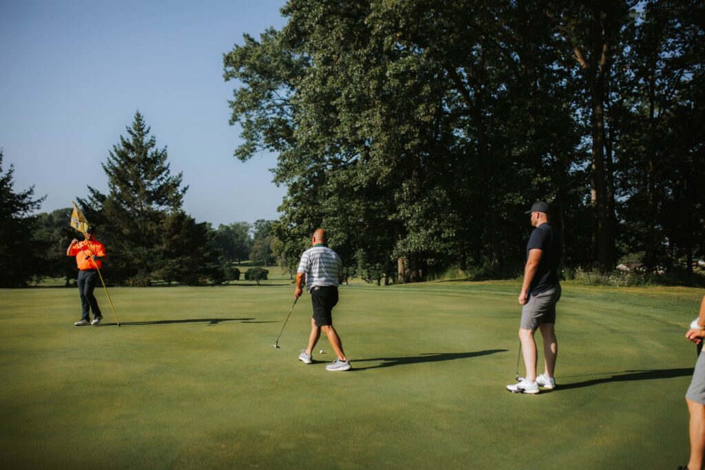 Three men stand on a golf green; one holds a flag, another prepares to putt, and the third watches. Tall trees surround the golf course under a clear blue sky.