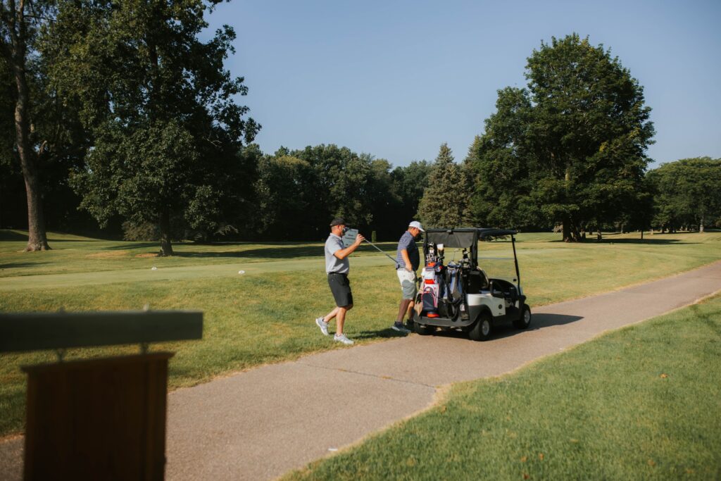 Two people stand beside a golf cart on a paved path at a golf course, surrounded by green grass and trees under a clear blue sky. One person holds a golf club, and golf bags are visible on the cart.