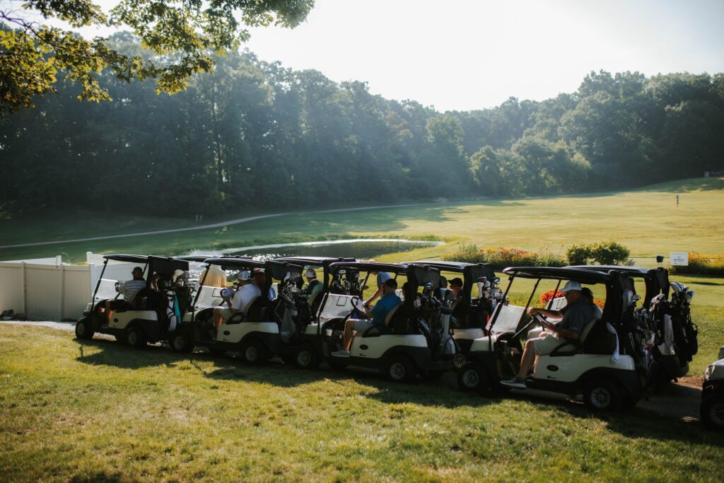 A row of people sitting in several golf carts, parked on grass beside a golf course on a sunny day, with trees and a winding path in the background.