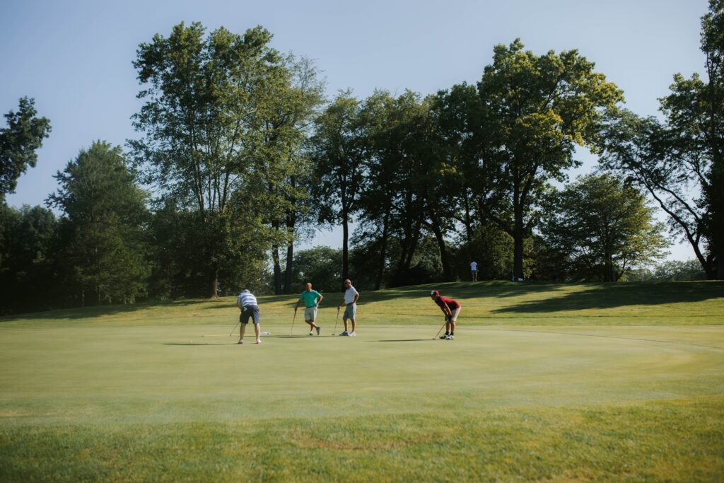 Four people are playing golf on a green course surrounded by trees on a sunny day. Three stand upright while one bends down, focusing on a putt. The background features lush, leafy trees and a clear sky.