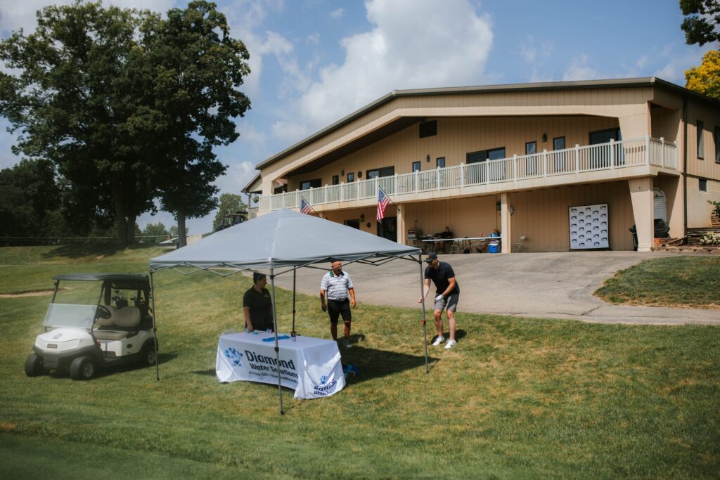 Three people stand under a canopy at a table with a “Diamond Healthcare” banner on a grassy area near a golf cart, in front of a large beige building with American flags and a patio.