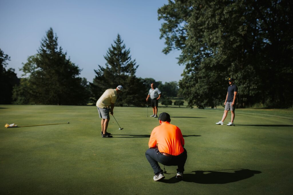 Four men are on a golf course; one is putting while another squats to observe. Two others stand in the background. Tall trees and a blue sky surround the green.