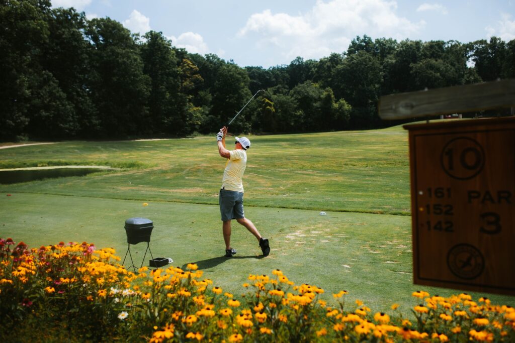 A golfer in a yellow shirt and shorts swings a club on a green golf course, surrounded by trees and yellow flowers, with a sign showing hole 10, par 3, and yardages in the foreground.