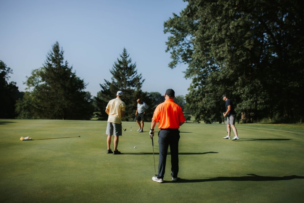 Four people stand on a grassy golf course on a sunny day, preparing to putt. Trees surround the green, and golf clubs and flags are visible. One person wears a bright orange shirt.