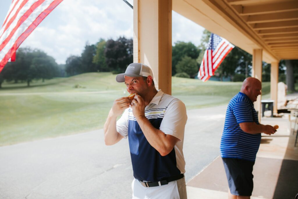 A man in a golf outfit and cap eats a sandwich outdoors near a golf course, with American flags hanging nearby. Another man in a striped shirt stands in the background. The weather is sunny with green grass visible.