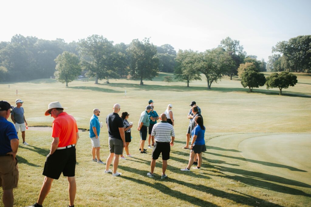 A group of people stand and chat on a grassy golf course near a putting green, with trees and blue sky in the background. Some hold golf clubs and wear hats, shorts, and polo shirts.