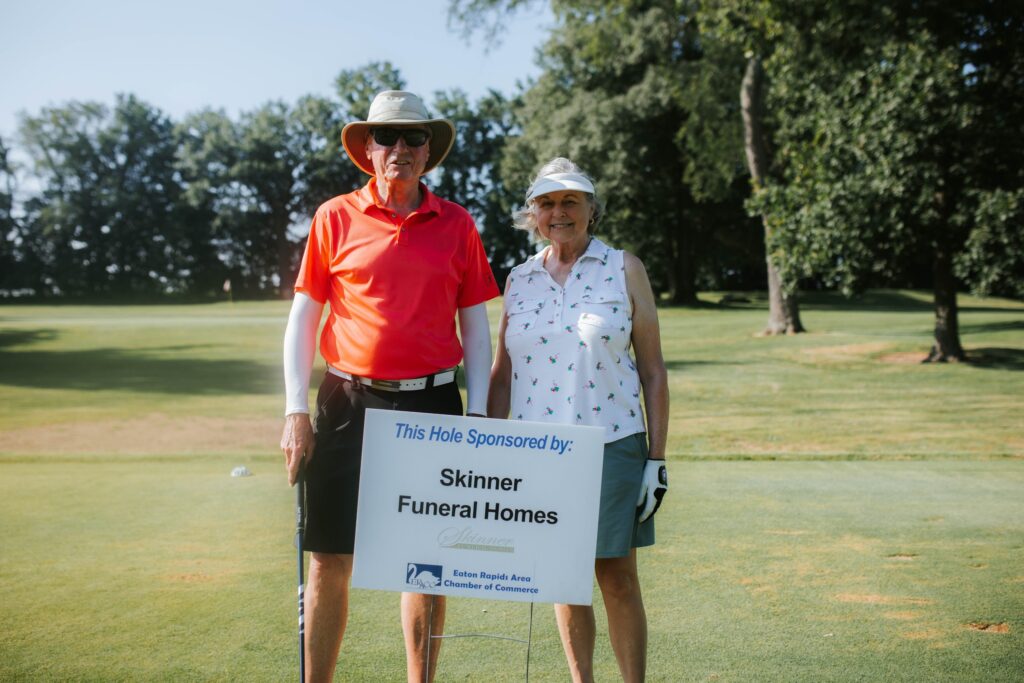 An older man and woman in golf attire stand on a golf course next to a sign that reads, This Hole Sponsored by: Skinner Funeral Homes. Trees and grass are visible in the background.