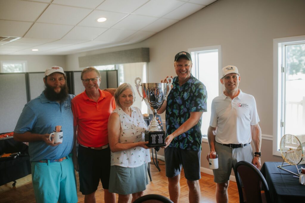Five people stand indoors, smiling at the camera. The central person holds a large trophy, while others hold mugs and a can. They appear to be celebrating an achievement, possibly at a golf or sports event.