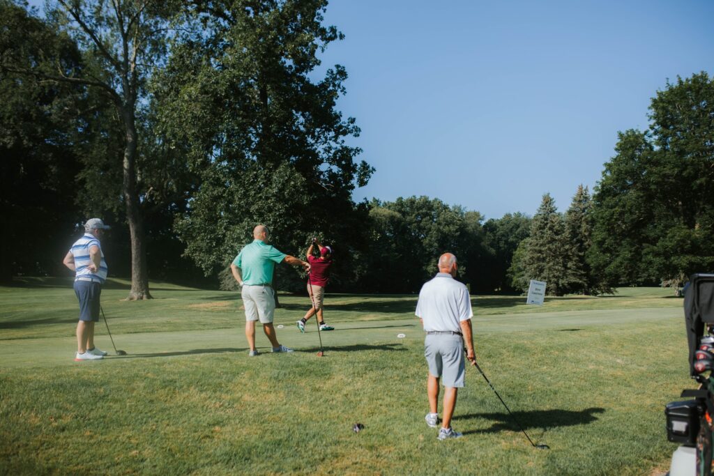 Four people are on a golf course; one person is swinging a golf club while the other three watch. Tall trees and green grass surround them under a clear blue sky.