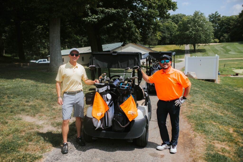 Two men stand next to a golf cart on a sunny day, each holding golf clubs. One man wears a yellow shirt and shorts, the other wears an orange shirt and dark pants. Trees and a golf course are visible in the background.