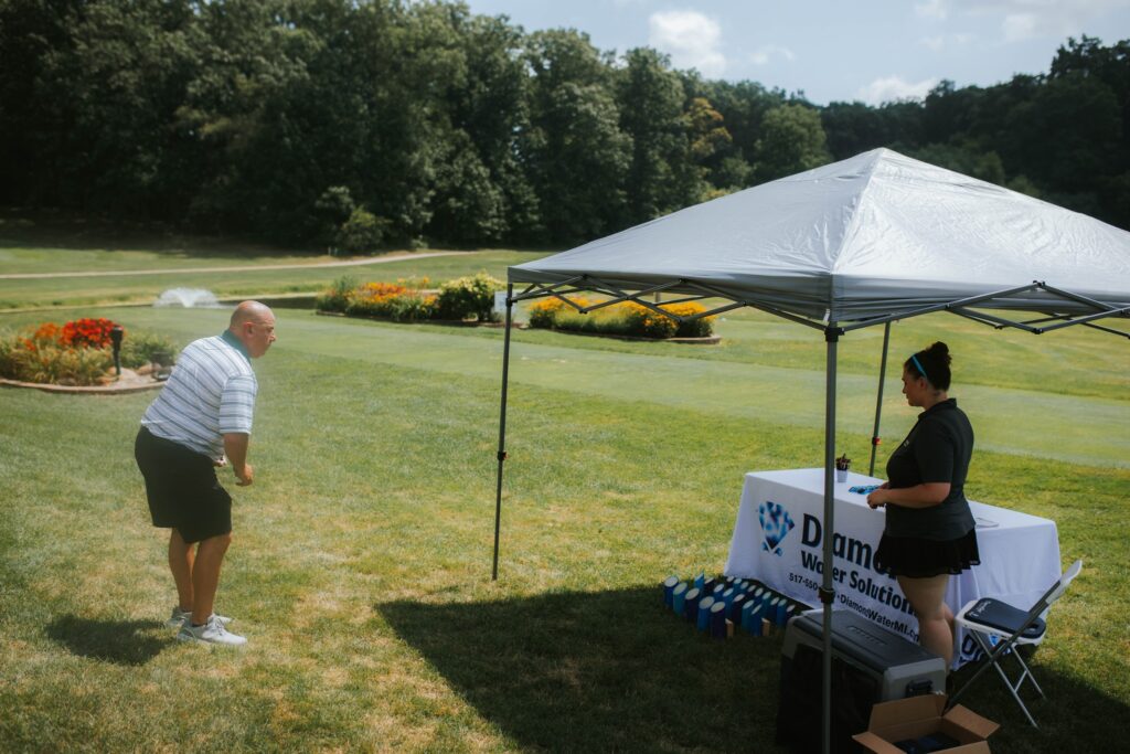 A man prepares to hit a golf ball on a green lawn near a tent, where a woman stands behind a table with promotional items and a Diamond Water Solutions banner. Trees and flower beds are in the background.