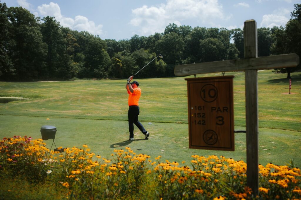 A golfer in an orange shirt swings a club on a green golf course near a wooden sign for hole 10, par 3, surrounded by yellow flowers and trees in the background under a blue sky.