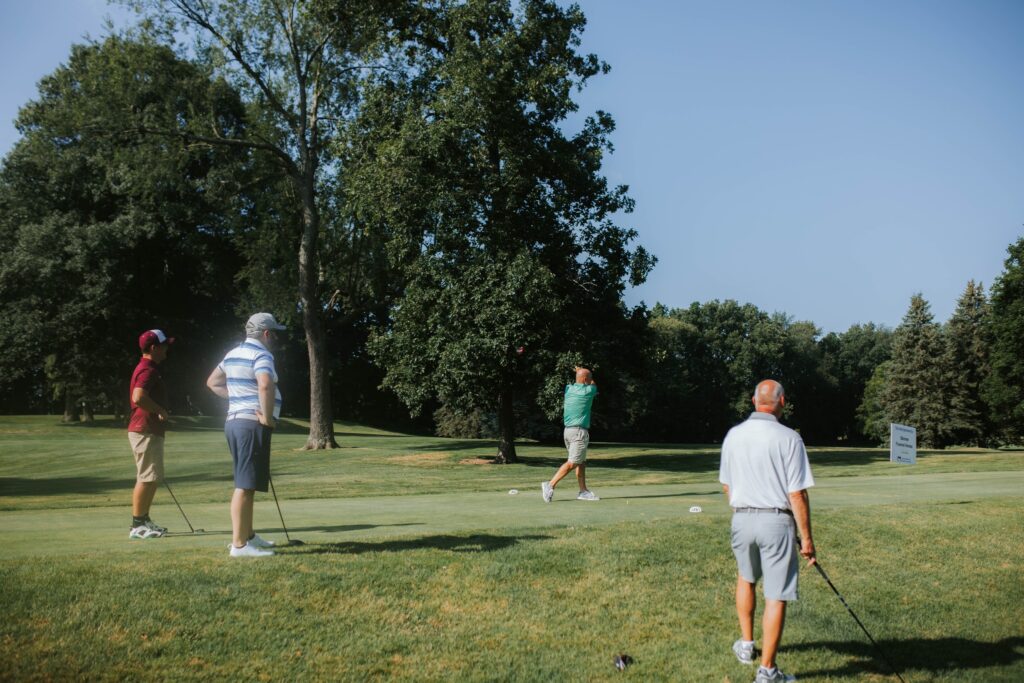 Four men stand on a green golf course, with one swinging a golf club while the other three watch. Tall trees and a blue sky are in the background.