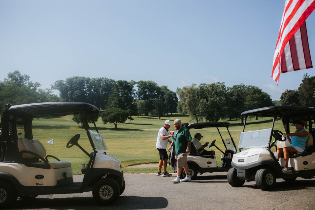 People gather near golf carts on a sunny day at a golf course, with green grass, trees, and a clear blue sky in the background. An American flag is visible on the right side of the image.