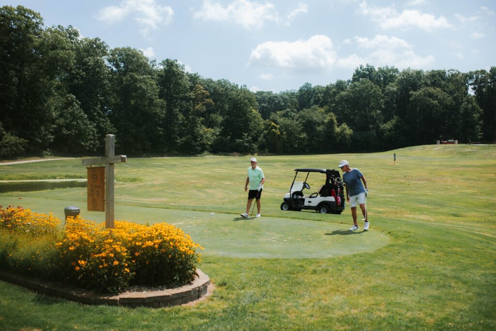 Two people stand on a golf course near a golf cart and a garden bed with yellow flowers. Trees line the background under a partly cloudy sky.